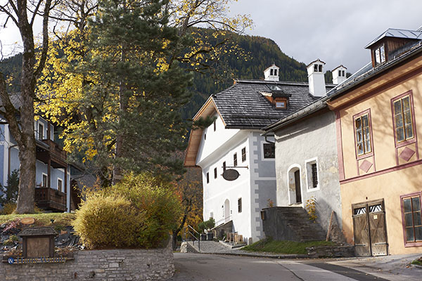 Foto vom Mesnerhaus sowie der unmittelbaren Umgebung (Wege, Nachbarhäuser, Bäume & Sträucher, Mauer, Hügel) im Herbst